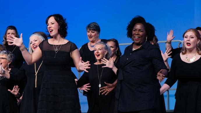 罗莎琳德·琼斯在她的全女子无伴奏合唱团中表演爵士手舞. 所有成员都在舞台上穿着黑色的衣服，背景是蓝色的