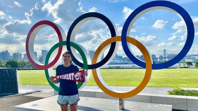 Brianna Tammaro stands in front of a statue of the Olympic rings holding a Trinity pennant