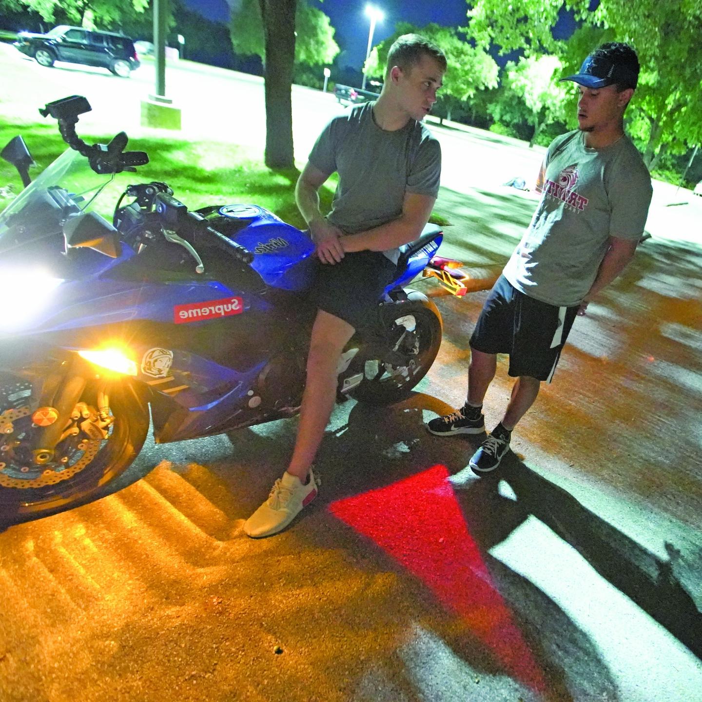 克里斯·斯图尔特和鲍比·马吉坐在一辆摩托车上，他们脚下的柏油路上投射着一个红色的转向灯.